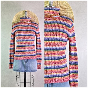 Pode incluir: Uma camisola de gola alta com mangas compridas e riscas horizontais multicoloridas. As riscas são vermelhas, laranja, amarelas, azuis e roxas. A camisola é exibida num manequim com um chapéu de palha e uma saia de ganga.