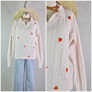 Peut inclure: Un pull rose clair en tricot torsadé avec un col et des fraises brodées. Le pull à manches longues est présenté sur un mannequin avec un jean bleu clair et un chapeau de paille.