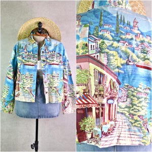 May include: A light blue denim jacket with a colourful coastal town print. The jacket has a button-up front, long sleeves, and a classic collar. The print shows buildings, boats, and a restaurant with a red and white striped awning.