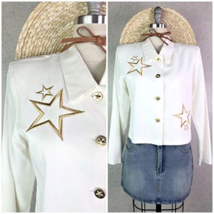 May include: White blazer with gold star embroidery and gold star-shaped buttons. The long-sleeved jacket is paired with a denim skirt and a straw hat. The blazer features a collared neckline and a tailored fit.