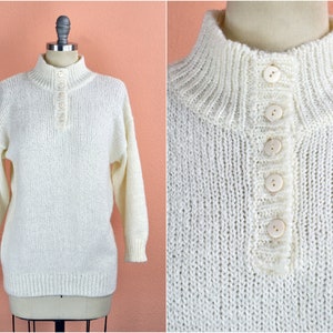 May include: A white knitted sweater with a mock neck and a button-up placket. The sweater has a textured knit and is made of a soft, cozy material.