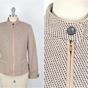 Puede incluir: Una chaqueta beige y negra con estampado, cierre de cremallera y cuello alto. La chaqueta tiene un ribete de cuero marrón alrededor del cuello y los bolsillos.