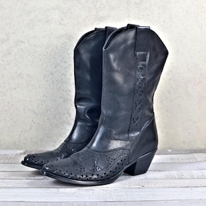 Könnte beinhalten: Ein Paar schwarze Leder-Cowgirl-Stiefel mit spitzer Zehenpartie und niedrigem Absatz. Die Stiefel haben ein dekoratives Ausschnittmuster und ein gesticktes Design am Schaft.