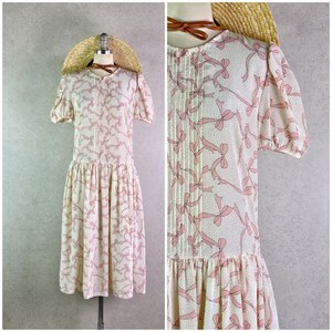 Könnte beinhalten: Cremefarbenes Vintage-Kleid mit rosa Schleifenmuster. Es hat kurze Ärmel, eine plissierte Vorderseite und einen gerafften Rock. Ein Strohhut mit braunem Lederband sitzt auf einem Schaufensterkopf, passend zum Kleid. Das Kleid ist ca. 100 cm lang.