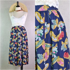 May include: A navy blue midi skirt featuring a pattern of yellow, pink, and green straw hats and floral details. The skirt is worn with a white lace top and a straw hat.