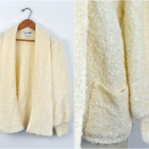 May include: A white fluffy cardigan with long sleeves and a shawl collar. The cardigan has a pocket on the left side.
