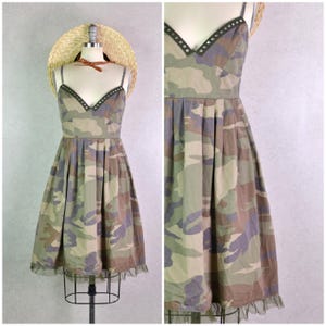 May include: A camouflage-print dress with a V-neck and thin straps. The dress has a fitted bodice, a pleated skirt, and a delicate ruffle at the hem. The neckline is embellished with small, dark studs. A straw hat with a brown ribbon sits on a mannequin head.