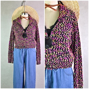 May include: A black, long-sleeved sweater with a V-neck and a colorful speckled pattern of pink, yellow, and green dots. The sweater has a black fur accent and is paired with blue jeans. A straw hat is in the background.