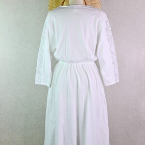 May include: A white dress with long sleeves and a gathered waist, displayed on a mannequin. The sleeves feature lace detailing. A straw hat rests on the mannequin's head, suggesting a summer or beach theme.