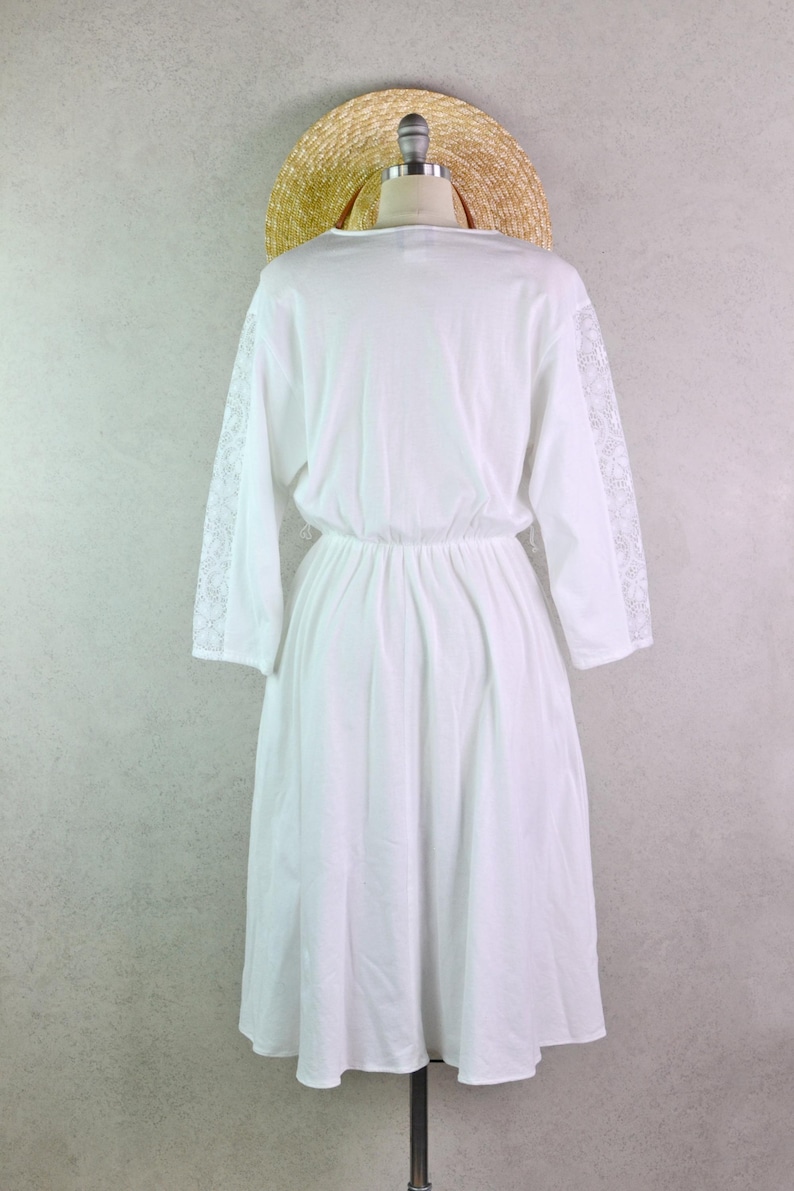 May include: A white dress with long sleeves and a gathered waist, displayed on a mannequin. The sleeves feature lace detailing. A straw hat rests on the mannequin's head, suggesting a summer or beach theme.