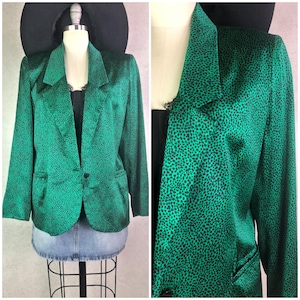 May include: A green blazer with a black spotted pattern. The blazer has a notched collar, two front pockets, and a single-button closure. The blazer is displayed on a mannequin with a black top and denim skirt.
