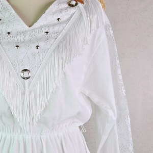 May include: White dress with a V-neckline, lace detailing, and fringe trim. The dress features long sleeves with lace accents and decorative silver-toned studs and buttons. A straw hat and brown leather strap are visible in the background.