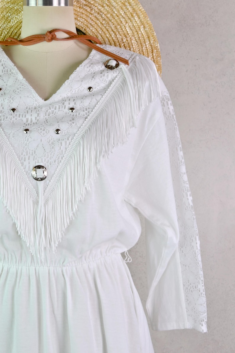 May include: White dress with a V-neckline, lace detailing, and fringe trim. The dress features long sleeves with lace accents and decorative silver-toned studs and buttons. A straw hat and brown leather strap are visible in the background.