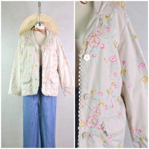 以下が含まれることがあります： ピンク、黄色、緑色の花の刺繍が施されたベージュのジャケット。 ジャケットには襟、ポケット、白いボタンが付いています。エッジは白いレースでトリミングされています。 ジャケットは麦わら帽子をかぶったマネキンに飾られています。