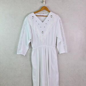 May include: White vintage dress with a V-neckline, fringe detail, and silver-tone embellishments. The dress has three-quarter sleeves with lace accents and a gathered waist. It hangs on a wooden hanger against a neutral background.