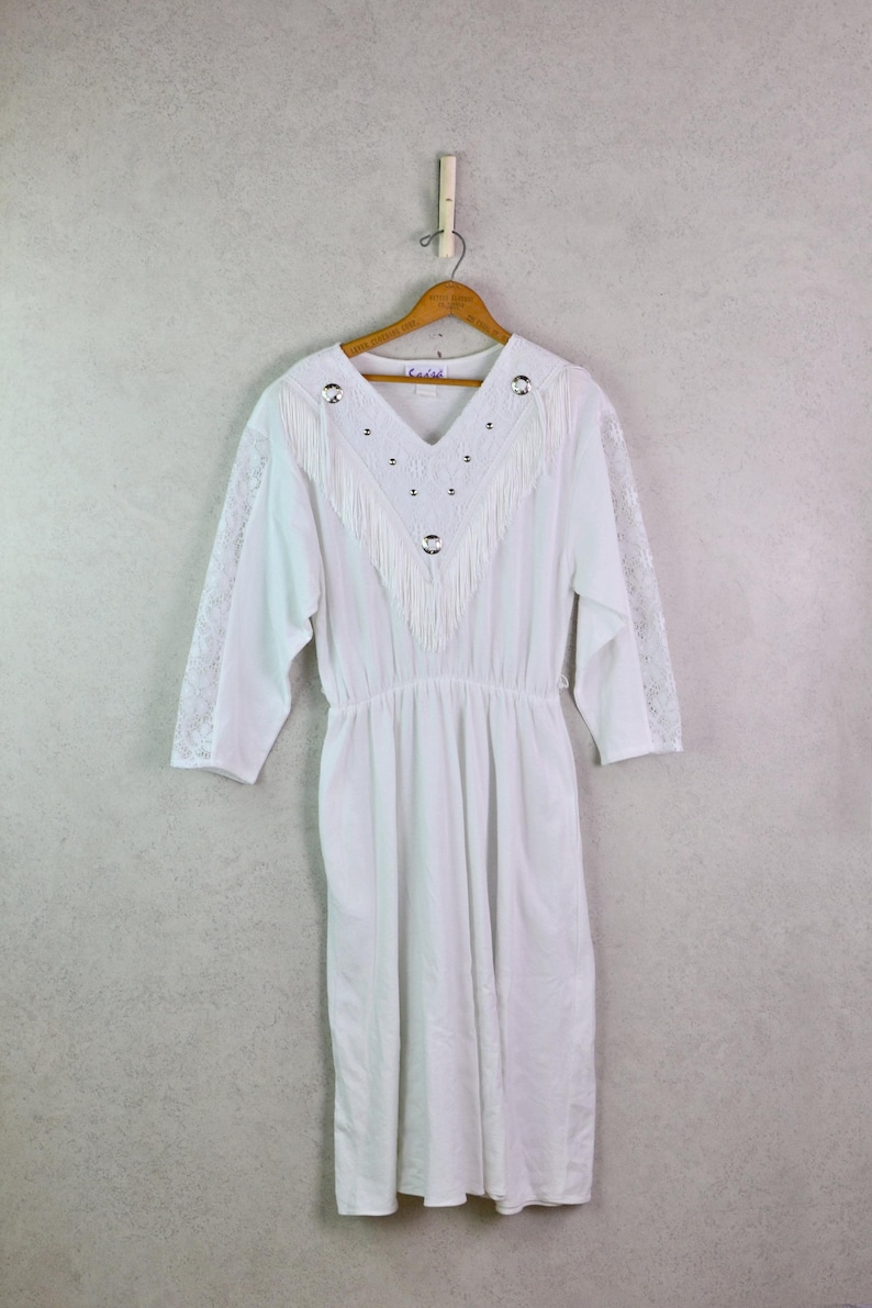 May include: White vintage dress with a V-neckline, fringe detail, and silver-tone embellishments. The dress has three-quarter sleeves with lace accents and a gathered waist. It hangs on a wooden hanger against a neutral background.