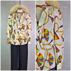 May include: A vintage white blouse with a colorful fan and ribbon pattern. The long-sleeved top features a wrap-style front and is paired with black pants. A straw hat and brown leather necklace are also visible.
