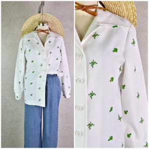 May include: White button-down shirt with long sleeves, featuring a pattern of green frog illustrations. The shirt is paired with blue denim pants. A straw hat and brown bow tie are also visible.
