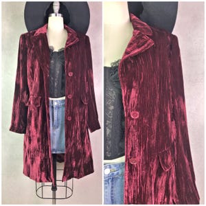May include: A long, burgundy velvet coat with a notched collar, two front pockets, and three buttons. The coat is displayed on a mannequin with a black top and denim skirt. A black hat sits atop the mannequin's head.