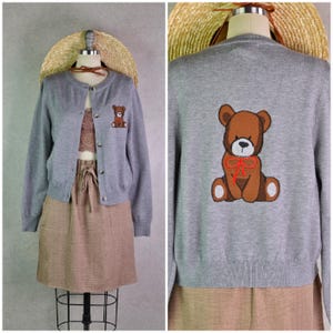 Può includere: Un cardigan grigio con un orsacchiotto marrone ricamato sulla tasca anteriore e sul retro. Il maglione ha uno scollo tondo e una chiusura con bottoni. L'orsacchiotto ha un fiocco rosso.
