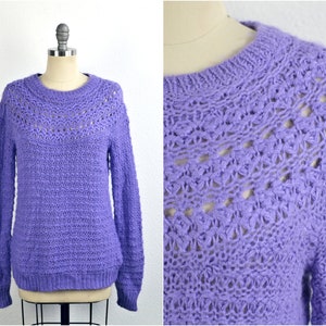 May include: A purple knitted sweater with a delicate openwork pattern around the neckline and shoulders. The sweater has a relaxed fit and long sleeves.