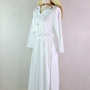 May include: A white dress with long sleeves and a V-neckline, adorned with fringe and silver studs. A straw hat with a brown leather band rests on the mannequin's head. The dress has an elastic waist and a flowing skirt.