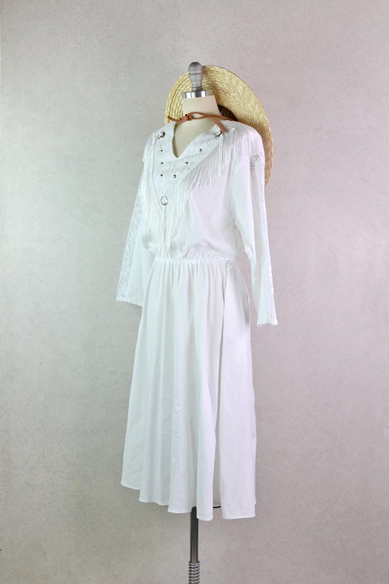 May include: A white dress with long sleeves and a V-neckline, adorned with fringe and silver studs. A straw hat with a brown leather band rests on the mannequin's head. The dress has an elastic waist and a flowing skirt.
