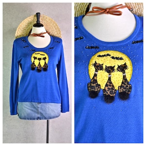 May include: A blue long-sleeve sweater featuring a whimsical Halloween design. The sweater showcases a yellow moon with three leopard-print cats, bats, and beaded accents. The sweater is displayed on a mannequin.