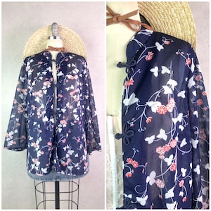 May include: Navy blue sheer jacket with a floral pattern in white and pink. The jacket has a mandarin collar and frog closures. A straw hat and brown leather strap are also visible.