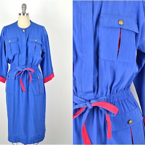 May include: A blue dress with a pink sash and gold buttons. The dress has long sleeves with pink cuffs and pockets on the front.