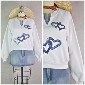 Op de afbeelding: Wit sweatshirt met lange mouwen en blauwe hartapplicaties. De V-hals en hartdetails hebben blauwe ruitjes. De manchetten en zoom zijn geribbeld. Een casual stijl.