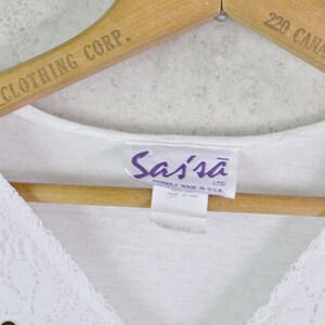 May include: A white V-neck top with lace detailing and a small silver embellishment. The top is hanging on a wooden hanger with the text "BETTER CLOTHES CO. 710358" and "CLOTHING CORP." printed on it. The tag reads "Sassa LTD. PROUDLY MADE IN USA."