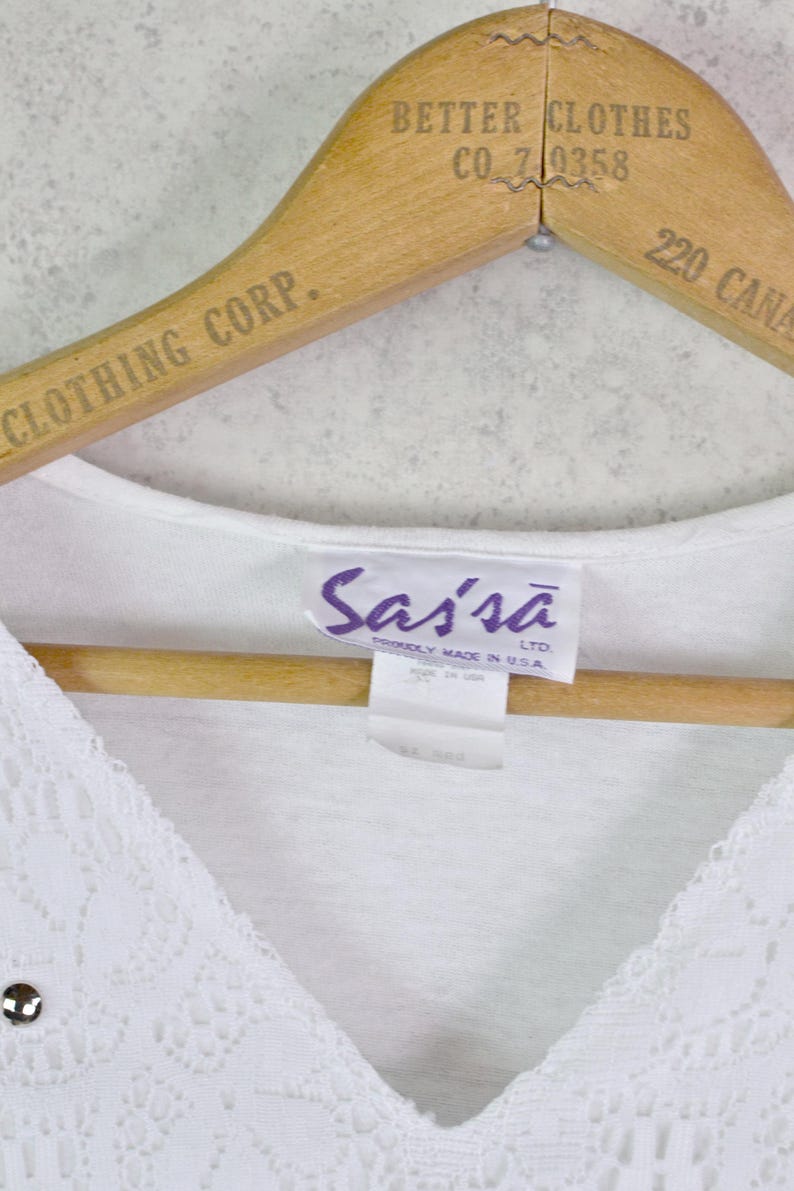 May include: A white V-neck top with lace detailing and a small silver embellishment. The top is hanging on a wooden hanger with the text "BETTER CLOTHES CO. 710358" and "CLOTHING CORP." printed on it. The tag reads "Sassa LTD. PROUDLY MADE IN USA."