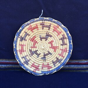 May include: A round woven basket with a blue, red, and white geometric pattern. The basket is made of natural materials and has a traditional design.
