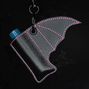 May include: A black leather case with a bat wing design, featuring pink stitching. The case holds a blue cylindrical object and is attached to a black metal clasp and chain. The background is solid black.