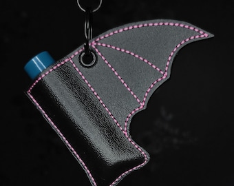 May include: A black leather case with a bat wing design, featuring pink stitching. The case holds a blue cylindrical object and is attached to a black metal clasp and chain. The background is solid black.
