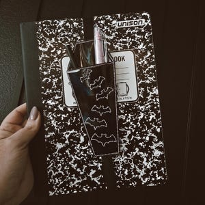 May include: A black and white composition notebook with a black pencil holder featuring a bat design. The pencil holder is attached to the notebook with a black strap. The notebook has the word "UNISON" printed on the cover.