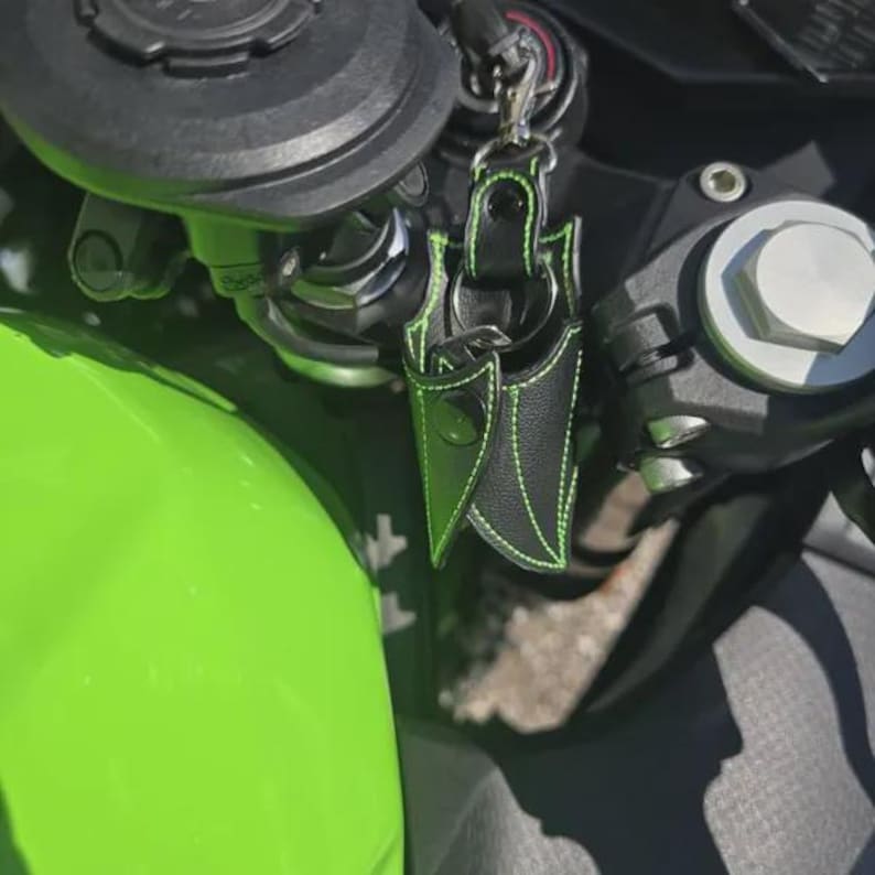 May include: A black leather key fob with bright green stitching, featuring a pointed, multi-layered design and a metal snap button closure, attached to a motorcycle handlebar assembly near a vibrant lime green fuel tank component.