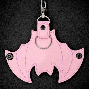 May include: A pink bat-shaped keychain with purple stitching and a silver keyring. The keychain has a metal clasp for easy attachment. The background is black.