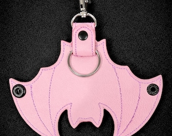 May include: A pink bat-shaped keychain with purple stitching and a silver keyring. The keychain has a metal clasp for easy attachment. The background is black.