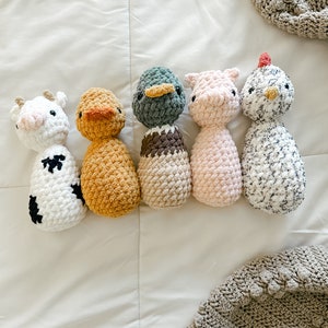 May include: A collection of handmade crochet farm animal toys, including a cow, duck, pig, and chicken. The cow is white with black spots, the duck is yellow, the pig is pink, and the chicken is white with black speckles. The toys are arranged on a white surface.