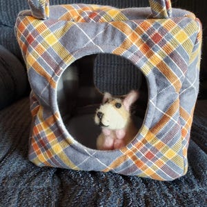 Puede incluir: Una pequeña cama cuadrada para mascotas con una abertura circular. La cama está hecha de tela gris con un estampado de cuadros en naranja, amarillo y marrón. Una pequeña figura de perro de fieltro está dentro.
