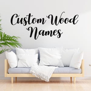 May include: A living room scene with a light wood-framed sofa, gray cushions, and white pillows. The text "Custom Wood Names" is written in black script on the white wall. A potted plant and a side table with a plant complete the decor.