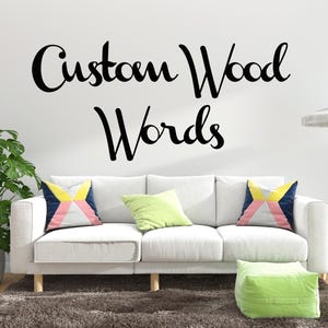 May include: A living room scene featuring a white sofa with colorful pillows and a green ottoman on a brown rug. The wall displays the text "Custom Wood Words" in black script. A potted plant and a side table with a lamp complete the decor.