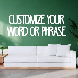 May include: A white sofa with two matching pillows sits in front of a green wall. The text "CUSTOMIZE YOUR WORD OR PHRASE" is displayed in white. Potted plants are on either side.