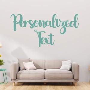 May include: A teal-colored wall decal with the words "Personalized Text" in a cursive font is displayed on a white wall. A beige sofa with two white pillows sits below the text. A side table with a plant and a lamp are also visible.