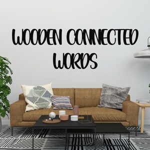 May include: A living room scene with a brown sofa, decorative pillows, and a black coffee table. The wall features the text "WOODEN CONNECTED WORDS" in a bold, black script. A large potted plant and a small side table with a plant are also visible.