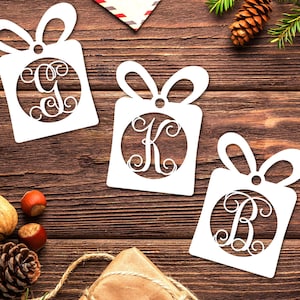 May include: Three white paper gift tag ornaments with a circle in the center and a single letter monogram in each circle. The letters are G, K, and B. The ornaments are on a brown wooden surface.