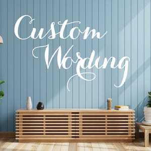 May include: A light blue wall with the words "Custom Wording" in white script. A wooden cabinet with horizontal slats sits below the text. Decorative items and plants are on the cabinet and in front of the wall.
