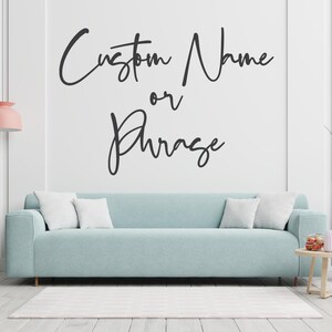 May include: A light blue sofa with white pillows sits in front of a white wall. The wall has the text "Custom Name or Phrase" in black script. A pink lamp and a small table with decor are also visible.